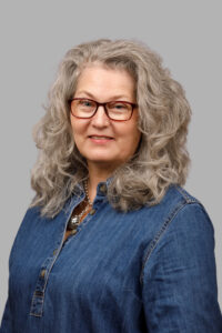 A headshot of a woman with wavy gray hair, red-framed glasses, and a denim shirt, smiling against a gray background.