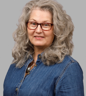 A headshot of a woman with wavy gray hair, red-framed glasses, and a denim shirt, smiling against a gray background.