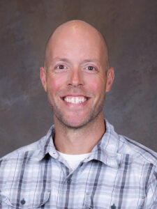 A smiling man with is wearing a light plaid button-up shirt. The background is a neutral, textured surface, making him the focal point of the image.