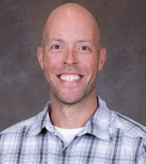 A smiling man with is wearing a light plaid button-up shirt. The background is a neutral, textured surface, making him the focal point of the image.