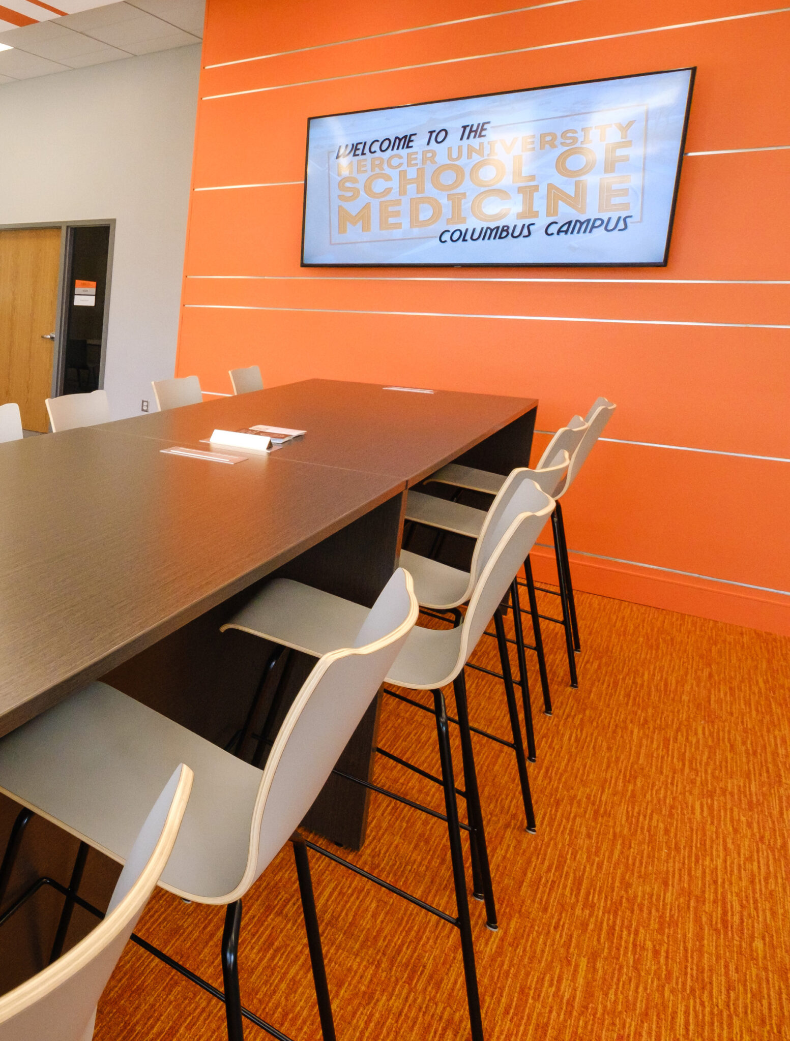 Table with chairs in front of orange wall with TV on itl