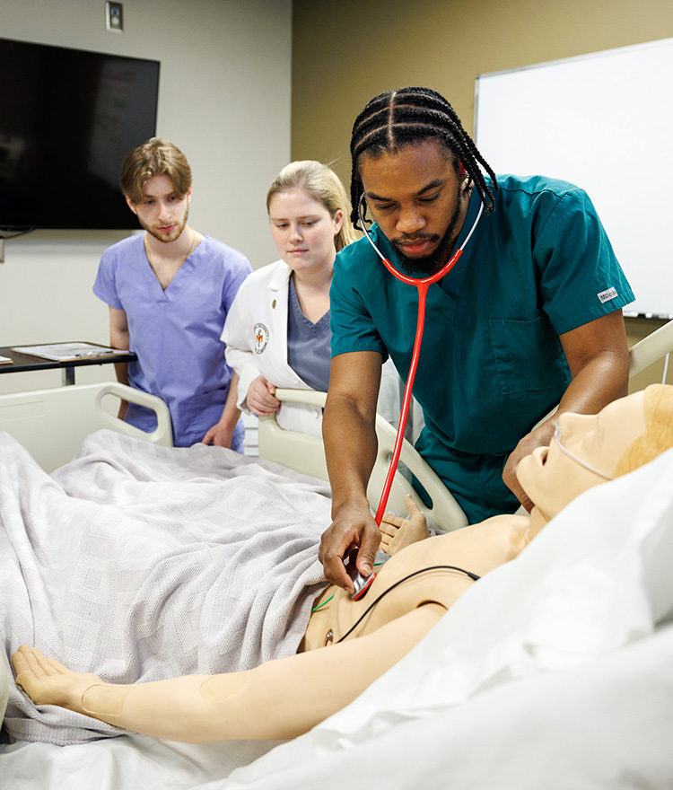 Three medical school students stand by full body medical mannequin.