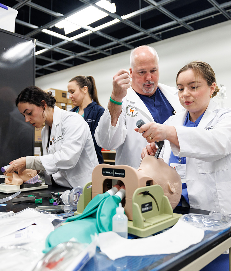 Two female students with white coats and one male instructor working on medical models.