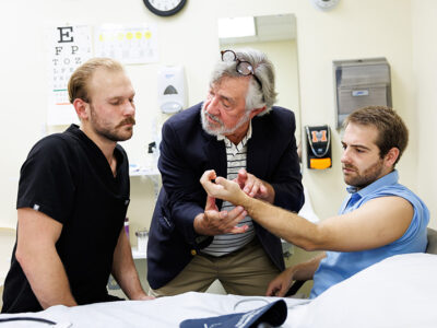 Older man checks the pulse of other young man while speaking to another male.