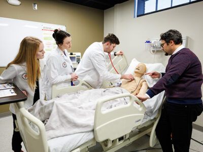 Threes students work on medical manikin while professor oversees.