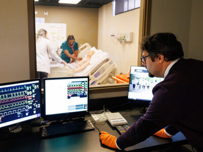 Man looks at computer screens through window of students working on medical manikin.
