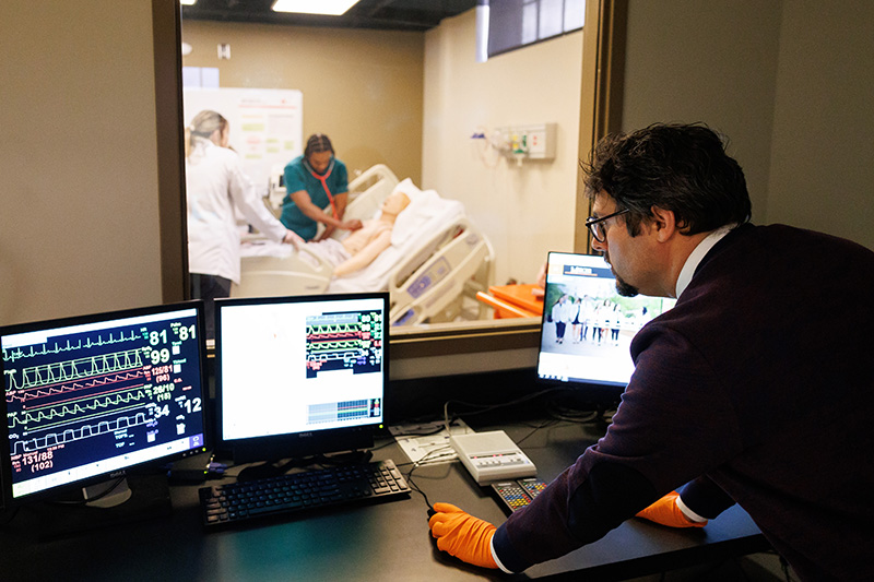 Man looks at computer screens through window of students working on medical manikin.