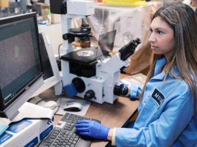 Female student in lab coat looking at slide on computer screen.