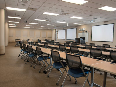 Large room with learning screens on one wall and many long tables with chairs.