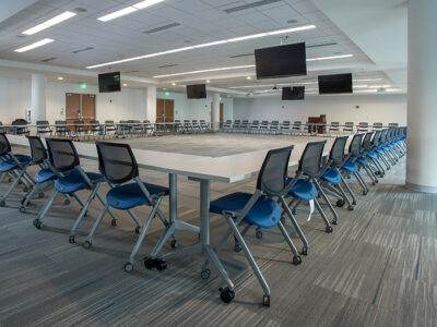 Large room with large monitors on walls and many long tables with chairs in the shape of a square.