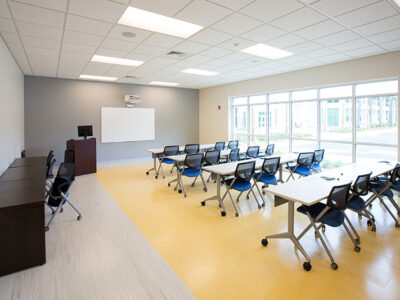 Large classroom with windows on one wall and may tables and chairs.