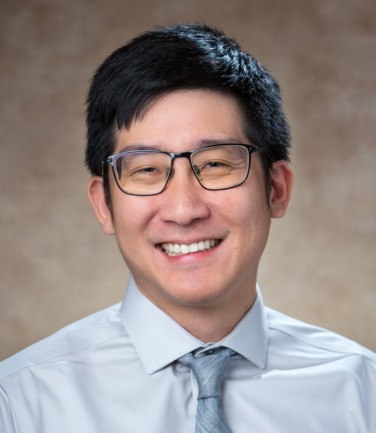 Dr. Tse-Yu Chen, wearing glasses, a light blue shirt and tie, smiles in front of a beige backdrop.