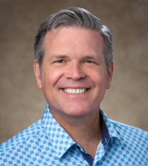 Dr. Jonathan Coffman, wearing a blue collared shirt, smiles in front of a beige backdrop.