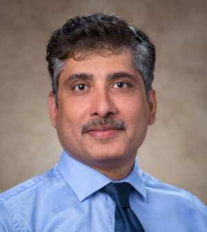 Dr. Sandeep Meharwal weasring a light blue shirt and dark blue tie, smiles in front of a beige backdrop