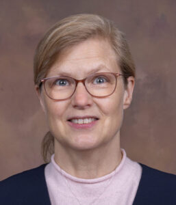 Dr. Andrea Winquist, wearing glasses and a pink shirt, smiles in front of a brown backdrop.