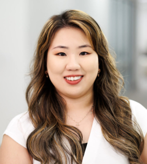 Woman with long, brown hair and smiling and wearing a white top in a bright, white hallway.