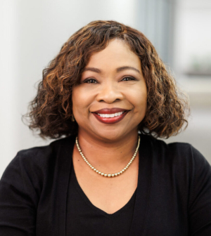 Woman with short, brown curly hair and smiling and wearing a black sweater in a bright, white hallway.