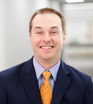 Dr. Jonathan Smith smiling wearing an orange tie and blue suit in a bright, white hallway